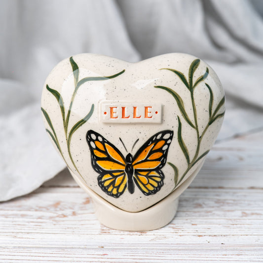Hand-painted heart-shaped ceramic urn with a butterfly and greenery on a stand for human or pet ashes.