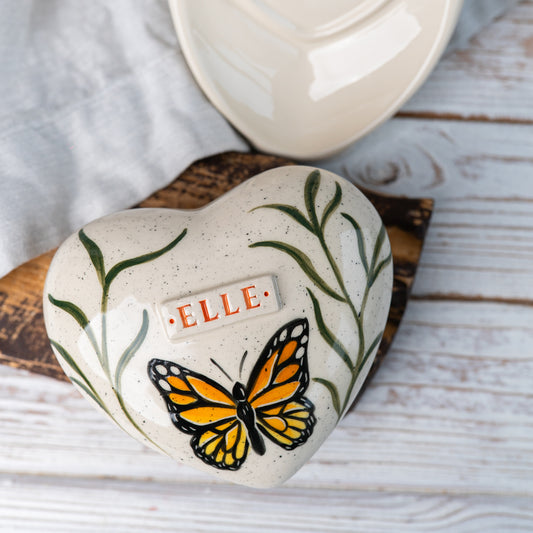 Top view of the heart-shaped urn showing the detailed butterfly wings and delicate green leaves.
