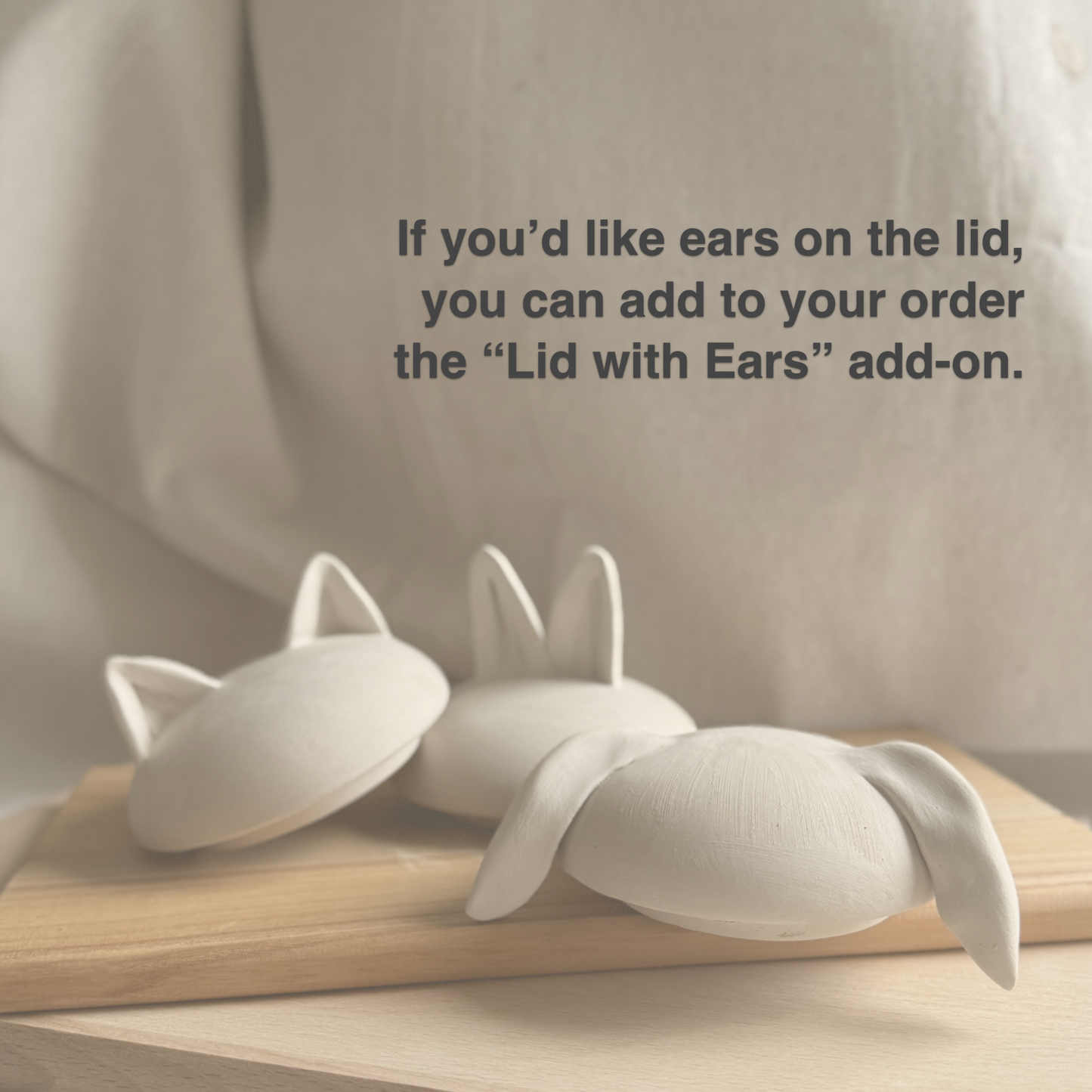 A photograph of three handmade, unglazed ceramic lids with custom clay ears on a wooden board. Dark grey text overlay in the upper right corner reads: 'If you'd like ears on the lid, you can add to your order the 'Lid with Ears' add-on.' The lids show three styles: small pointed cat ears (left), standing bunny ears (center), and long floppy bunny ears (right). The background is a soft linen fabric. This is an information slide detail the custom lid with ears add-on option for ceramic pet urns 