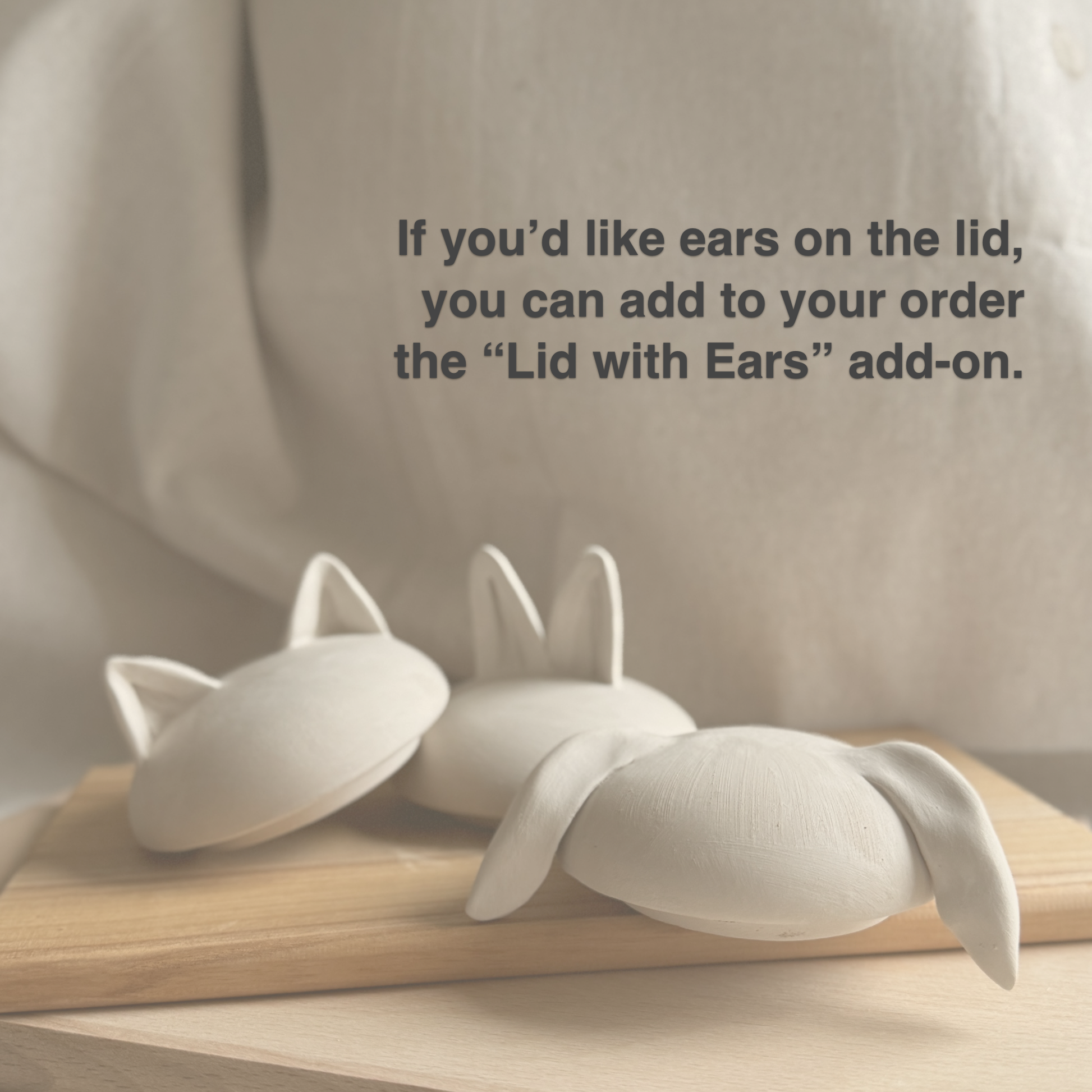 A photograph of three handmade, unglazed ceramic lids with custom clay ears on a wooden board. Dark grey text overlay in the upper right corner reads: 'If you'd like ears on the lid, you can add to your order the 'Lid with Ears' add-on.' The lids show three styles: small pointed cat ears (left), standing bunny ears (center), and long floppy bunny ears (right). The background is a soft linen fabric. This is an information slide detail the custom lid with ears add-on option for ceramic pet urns 