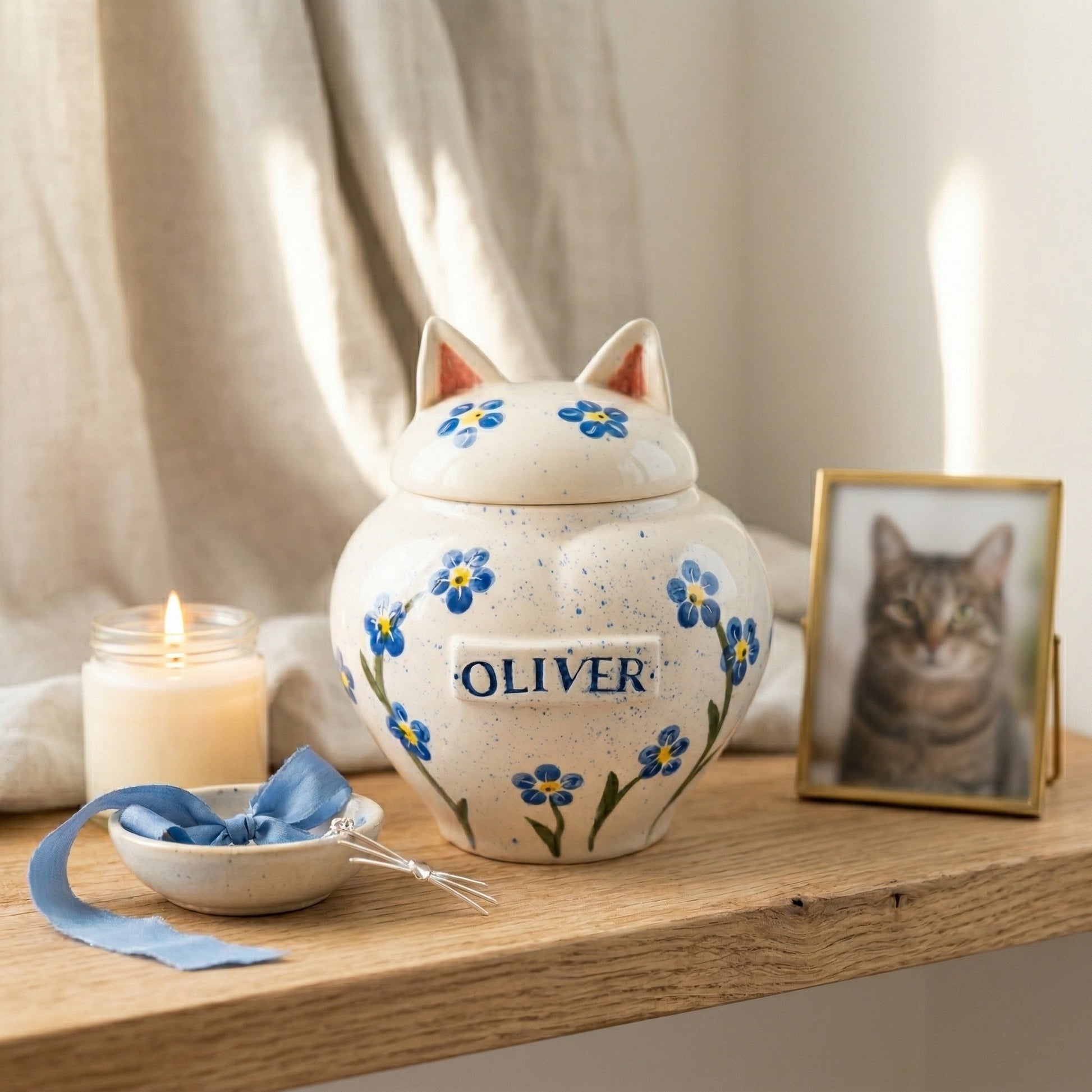 A serene home memorial display with a forget-me-not cat ceramic heart urn on a wooden shelf next to a small candle.