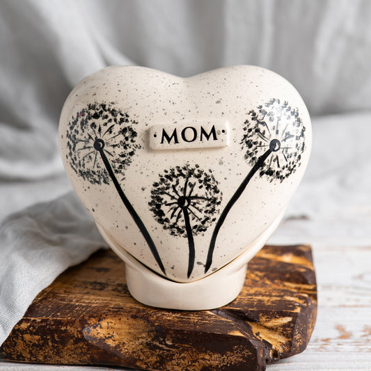 Hand-painted heart-shaped ceramic urn with a dandelion seed head and flying seeds on a stand.