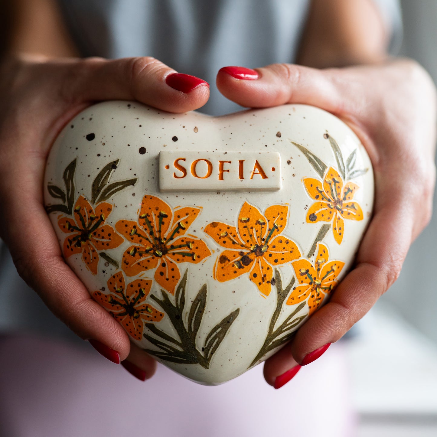 Holding a heart-shaped orange lily keepsake urn in hands to demonstrate the scale and hand-painted floral details.