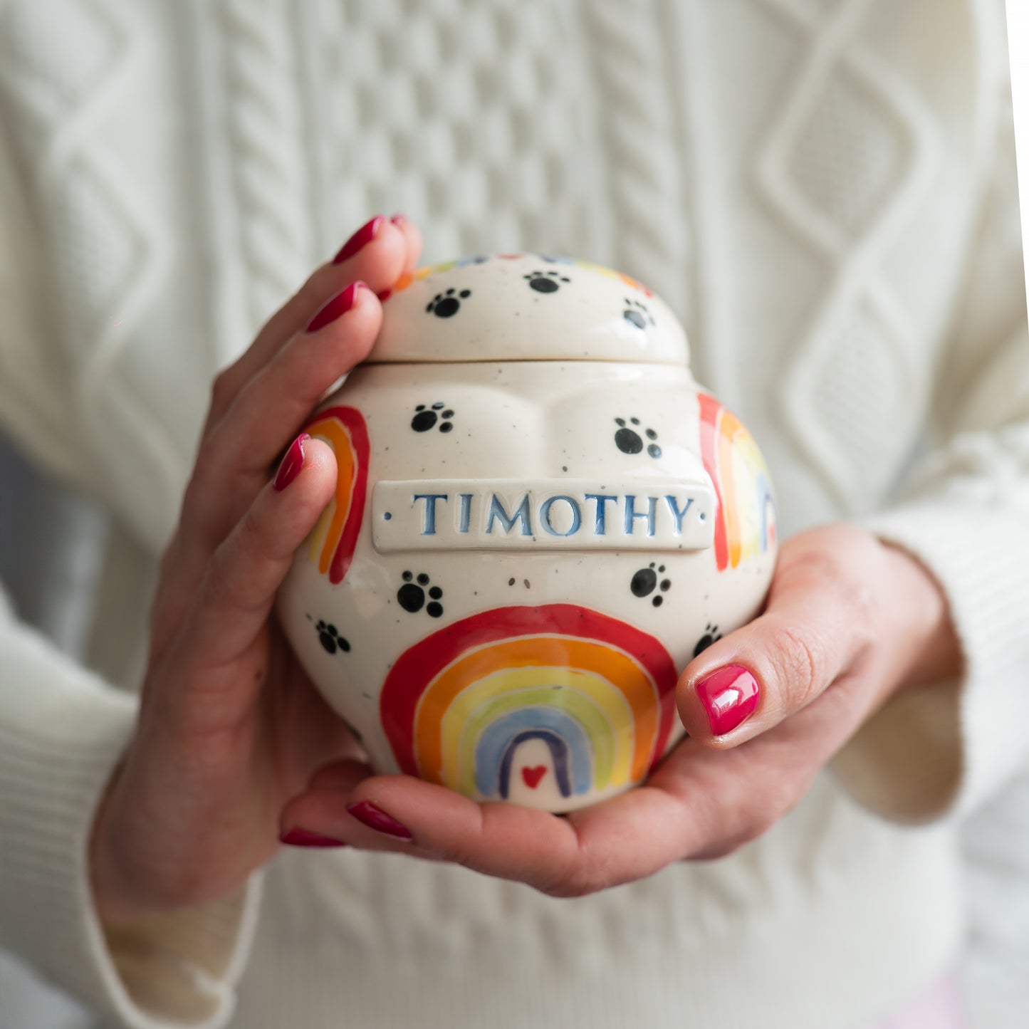 Holding a heart-shaped pot-style pet keepsake urn with a rainbow design in hands to show the scale and artisan detail.
