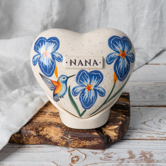 Hand-painted heart-shaped ceramic urn with blue irises and a hummingbird on a stand for human or pet ashes.