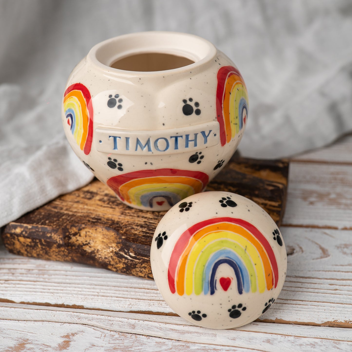 A close-up photograph of the hand-painted Rainbow Bridge pot-style pet heart ceramic urn with the lid removed. The fitted ceramic lid sits beside the open vessel, revealing the glazed interior designed to hold ashes.
