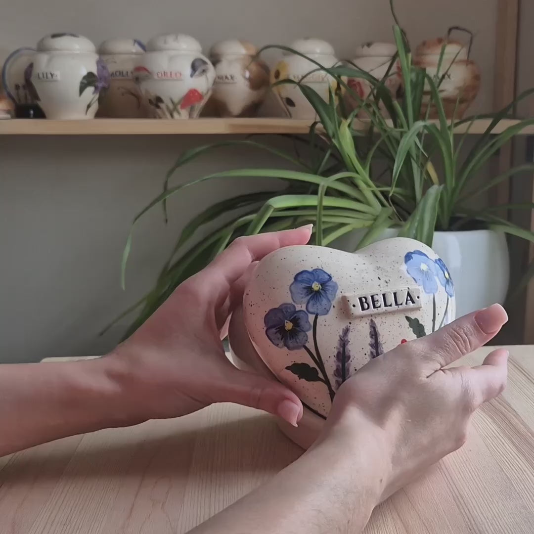 A close-up video demonstration of a hand-painted multi-flower heart-shaped ceramic keepsake urn. The video shows the urn being turned to display the vibrant pansies, lavender, and tiny flowers, along with the smooth glaze and matching stand.