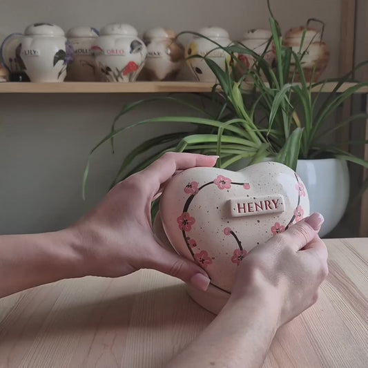 A close-up video demonstration of a hand-painted cherry blossom heart-shaped ceramic keepsake urn. The video shows the urn being held and slowly turned in hands to display the soft pink floral art, the smooth glossy glaze, and the matching custom ceramic stand.