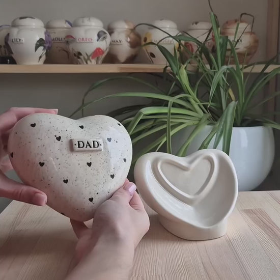 A close-up video demonstration of a hand-painted black hearts ceramic heart urn. The video shows the urn being turned in hands to display the minimalist design, the smooth glaze, and the matching stand.