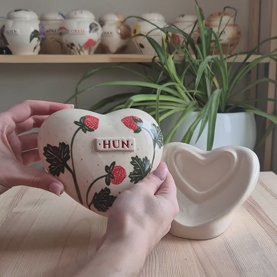A close-up video demonstration of a hand-painted strawberry heart-shaped ceramic keepsake urn. The video shows the urn being turned to display the vibrant berries, the smooth glaze, and the matching stand.