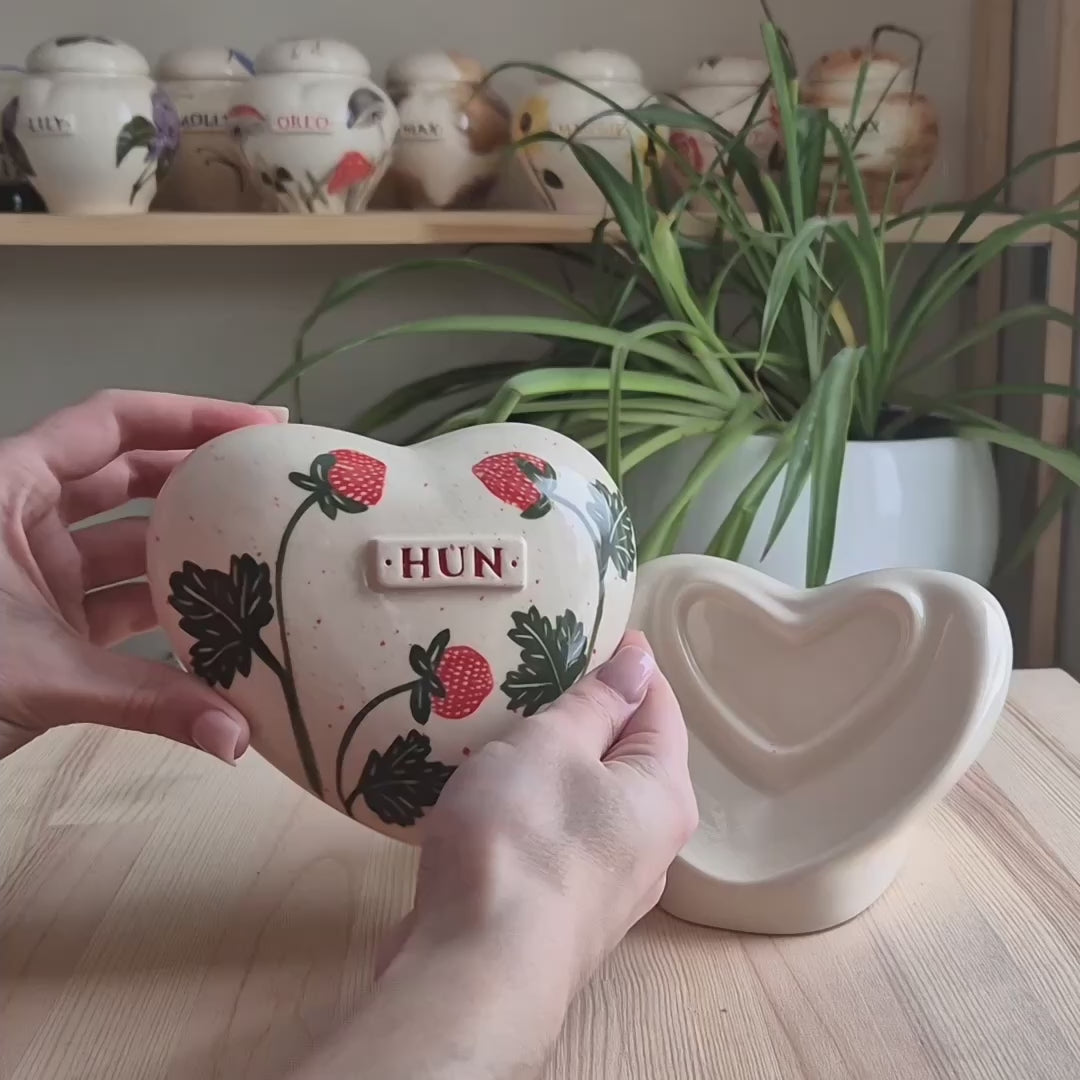 A close-up video demonstration of a hand-painted strawberry heart-shaped ceramic keepsake urn. The video shows the urn being turned to display the vibrant berries, the smooth glaze, and the matching stand.