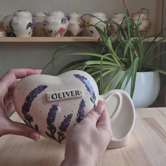 A close-up video demonstration of a hand-painted lavender heart-shaped ceramic keepsake urn. The video shows the urn being held and slowly turned in hands to display the delicate sprigs of lavender design, the smooth glazed finish, the snug-fitting lid, and the matching ceramic stand for display.