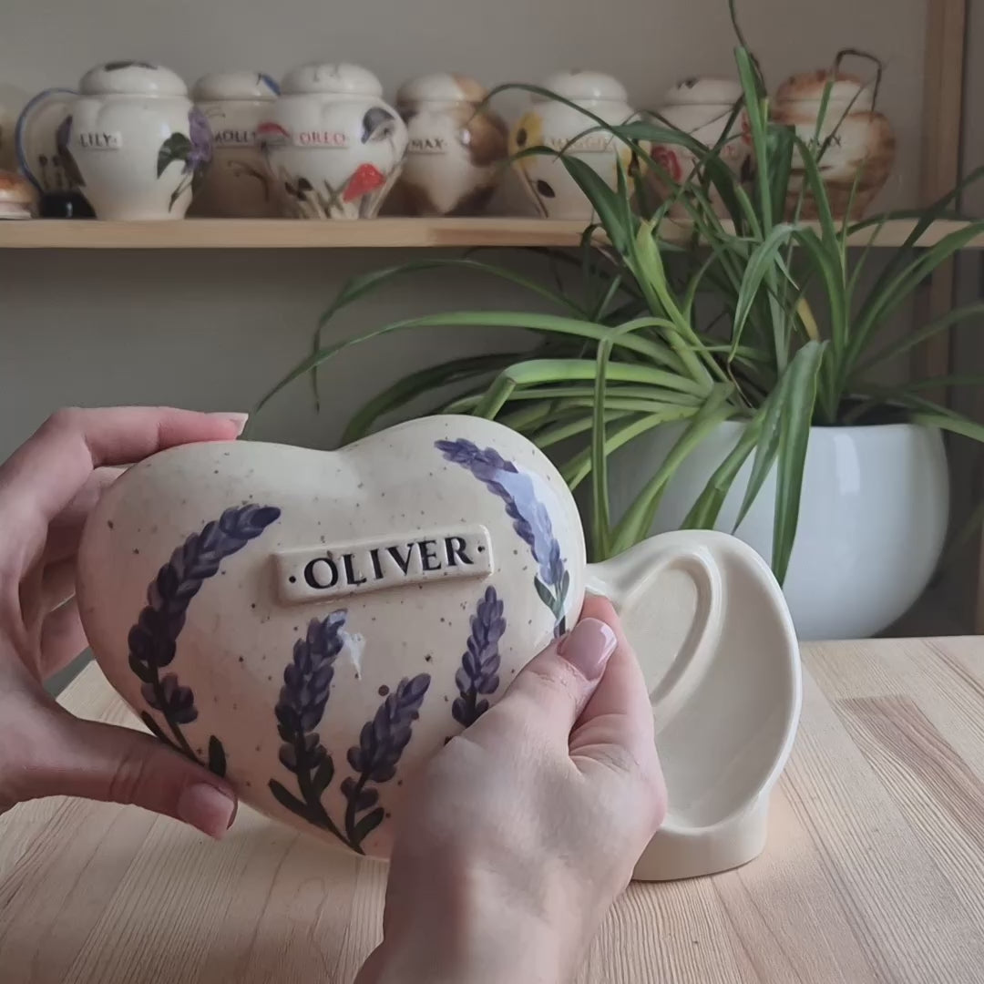 A close-up video demonstration of a hand-painted lavender heart-shaped ceramic keepsake urn. The video shows the urn being held and slowly turned in hands to display the delicate sprigs of lavender design, the smooth glazed finish, the snug-fitting lid, and the matching ceramic stand for display.