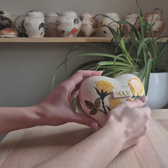 A close-up video demonstration of a hand-painted yellow rose heart-shaped ceramic keepsake urn. The video shows the urn being held and slowly turned to display the golden floral art, the smooth glaze, and the matching stand.