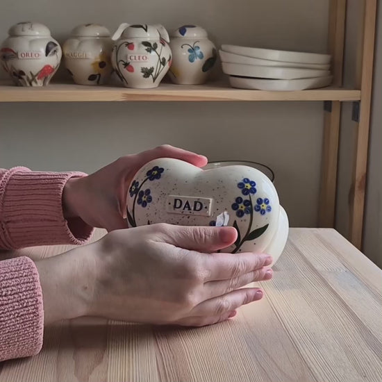A close-up video demonstration of a hand-painted forget-me-not heart-shaped ceramic keepsake urn. The video shows the urn being held and slowly turned in hands to display the delicate blue floral art, the smooth glossy glaze, and the matching ceramic stand.
