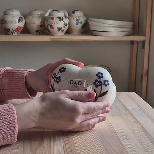A close-up video demonstration of a hand-painted forget-me-not heart-shaped ceramic keepsake urn. The video shows the urn being held and slowly turned in hands to display the delicate blue floral art, the smooth glossy glaze, and the matching ceramic stand.