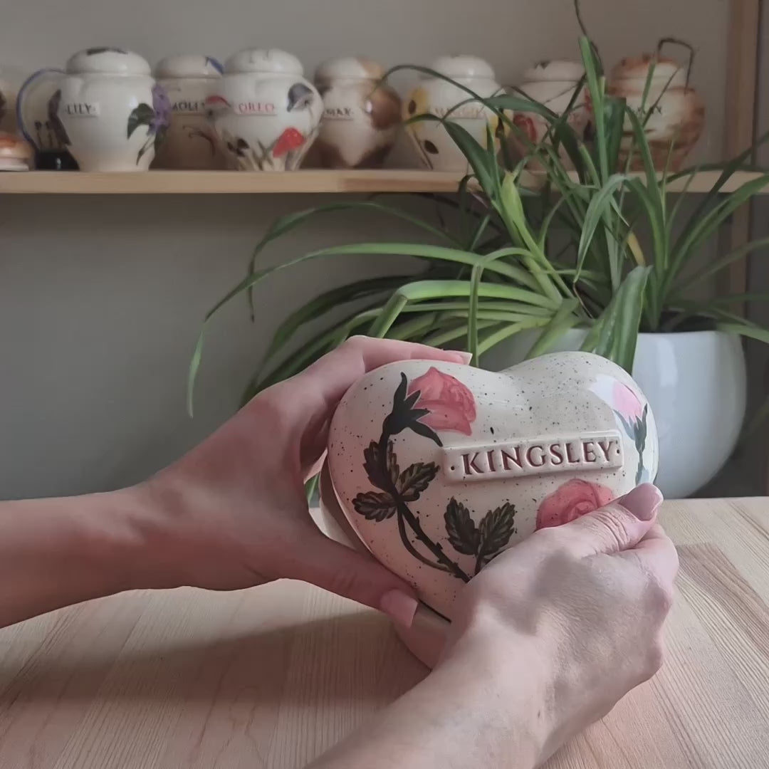 A close-up video demonstration of a hand-painted rose flower heart-shaped ceramic keepsake urn. The video shows the urn being held and slowly turned in hands to display the delicate floral details, the smooth glazed finish, the secure lid, and the matching ceramic stand for display.