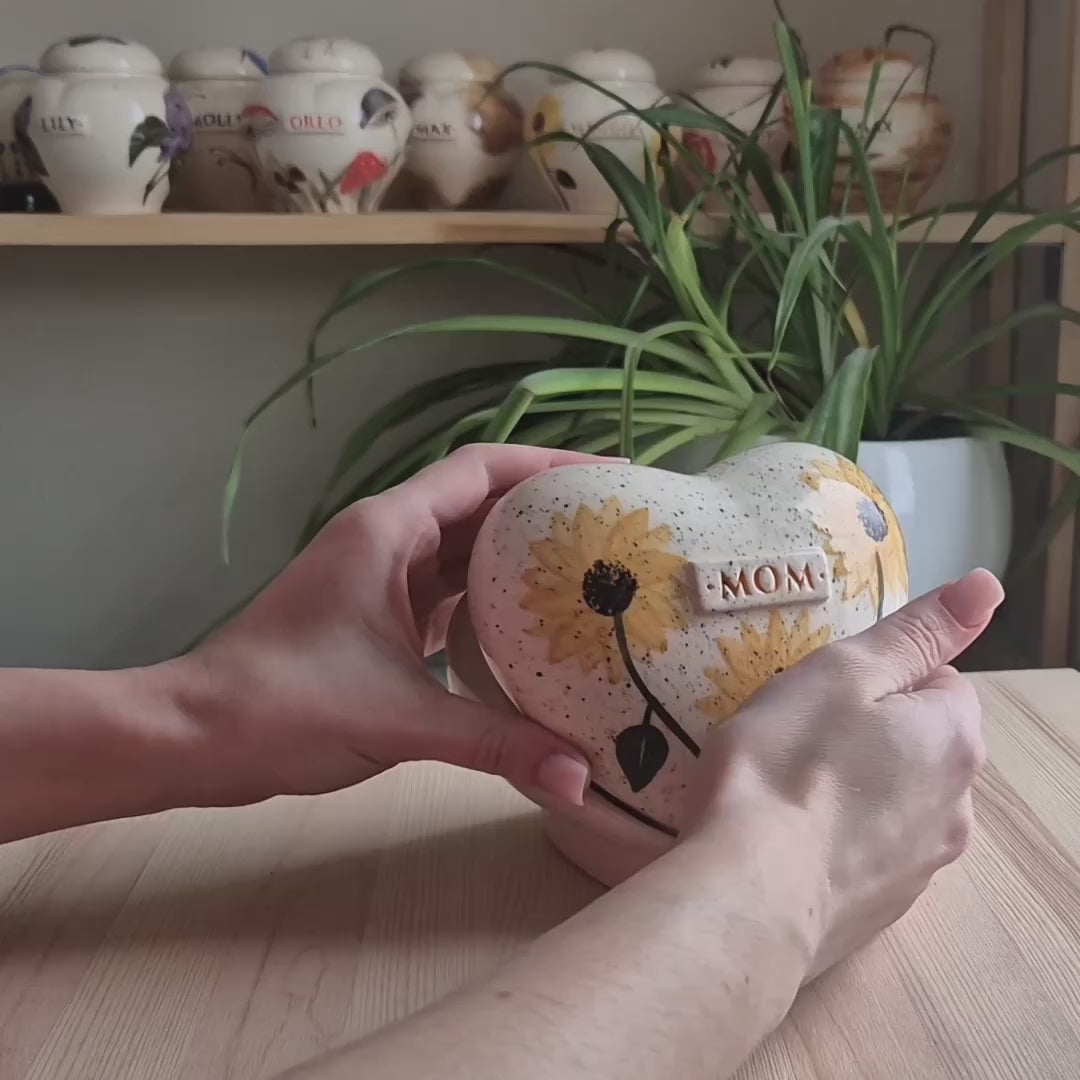 A close-up video demonstration of a hand-painted sunflower heart-shaped ceramic keepsake urn. The video shows the urn being held and slowly turned in hands to display the bright floral art, the smooth glaze, and the matching stand.