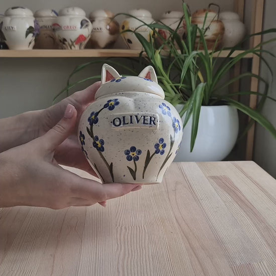 The video highlights the delicate blue flower patterns, the glossy white glaze, and the unique sculpted cat ears on the removable lid. It demonstrates the artisan texture and shows the lid being gently lifted to reveal the interior of this personalized memorial keepsake.